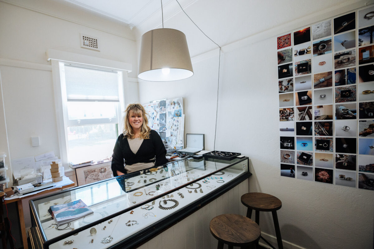 Michelle sits on a stool behind a glass viewing cabinet that is full for jewellery that she's made.