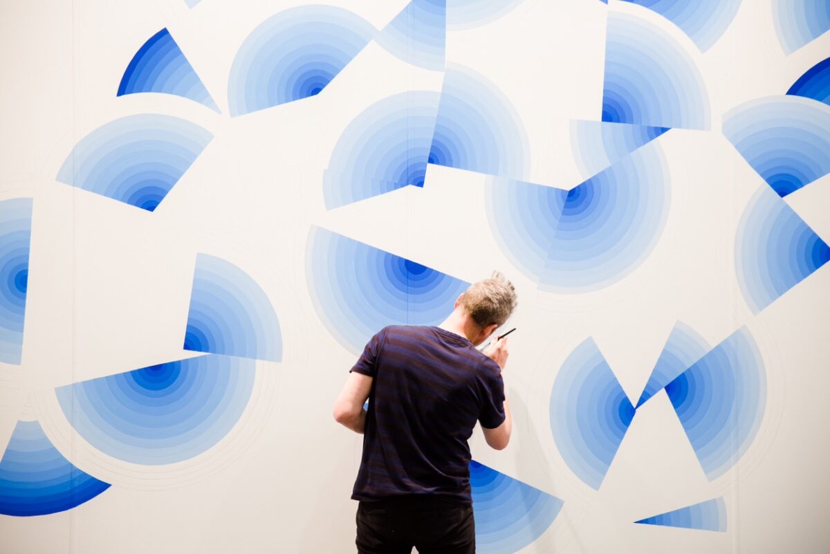 Photo of a man painting blue shapes on a large white wall.