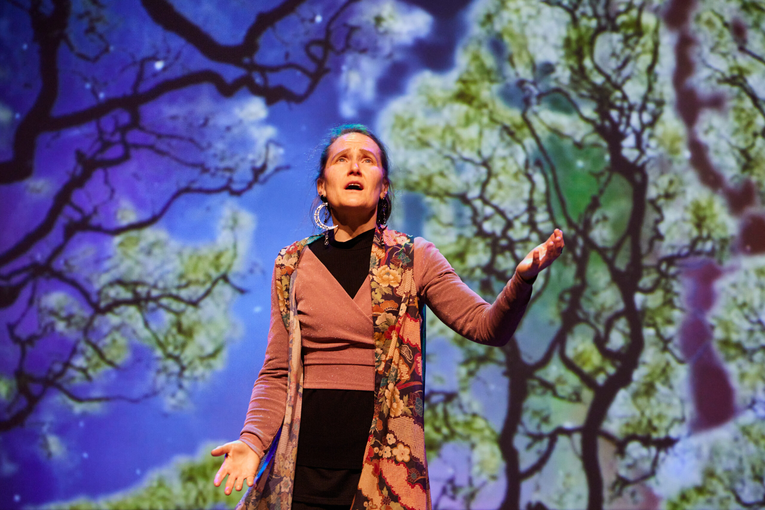 A woman dressed in earthy tones of brown and orange gestures on stage with a background projection of blue sky and green trees overhead.