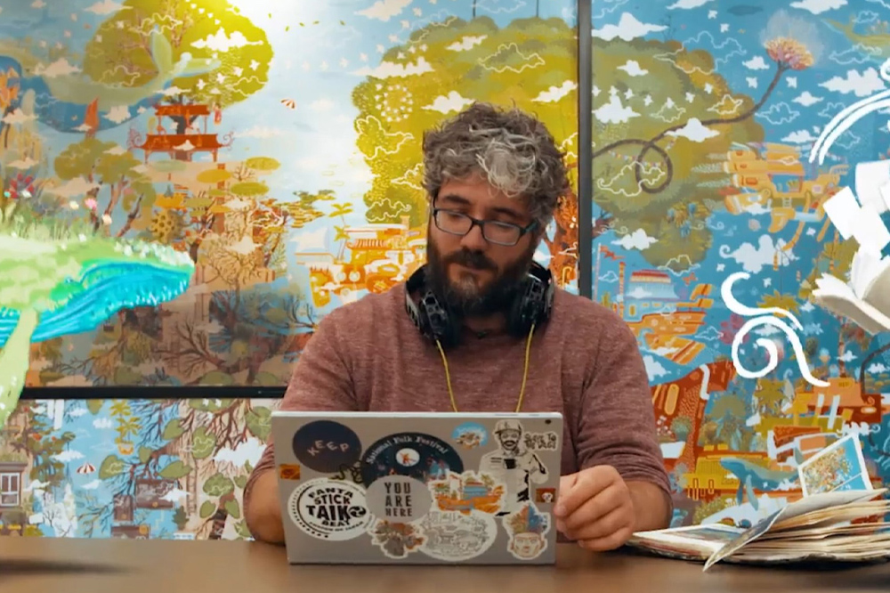 Paul sits in front of a big colourful mural that he created. He is working on his laptop which is covered in stickers
