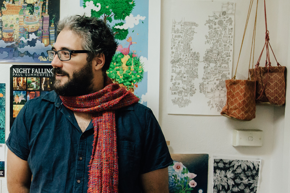 Paul wears a red scarf and black framed glasses and looks off to the side of the frame. Background is a collage of his colourful artworks.