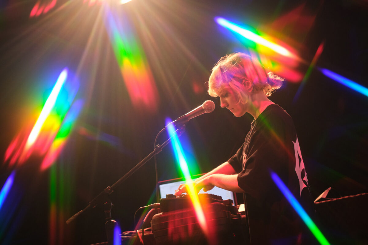 DJ with blond hair stands on the stage with rainbow lights around her.