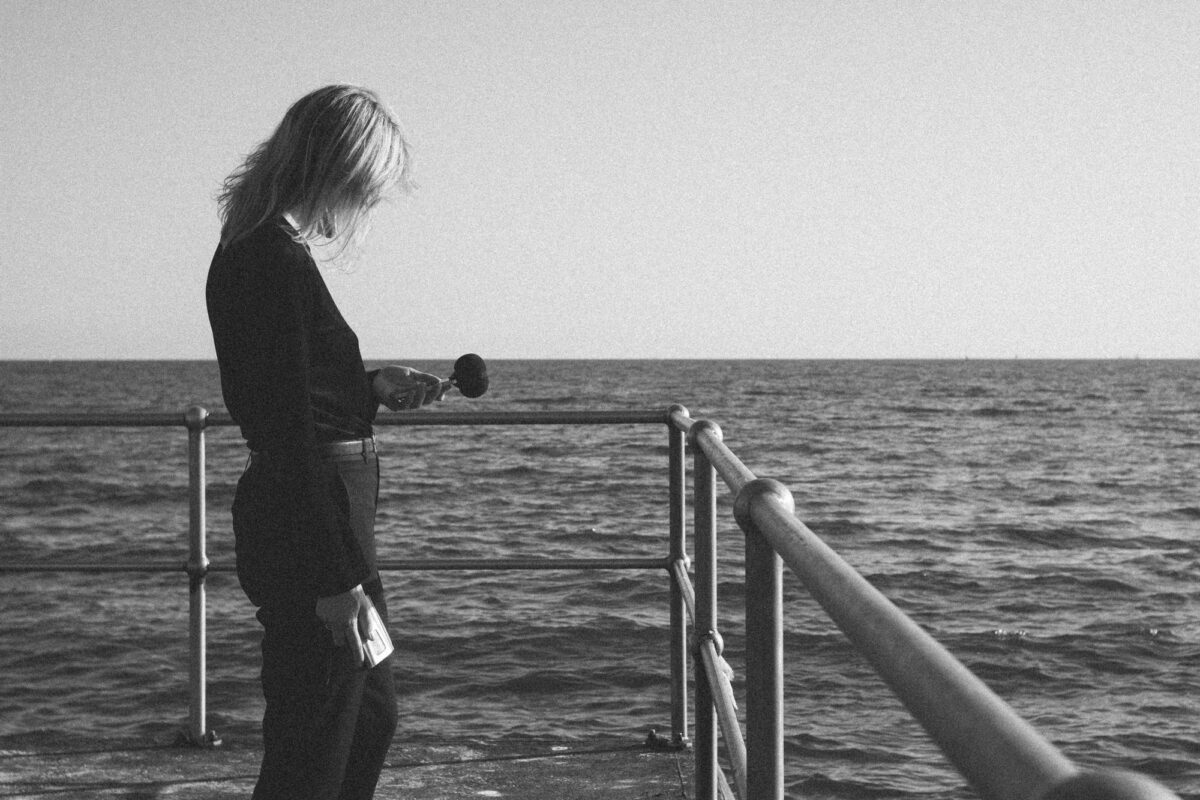 Black and White photo of a woman stands on a pier overlooking the sea. Promo for Ainslie Salon