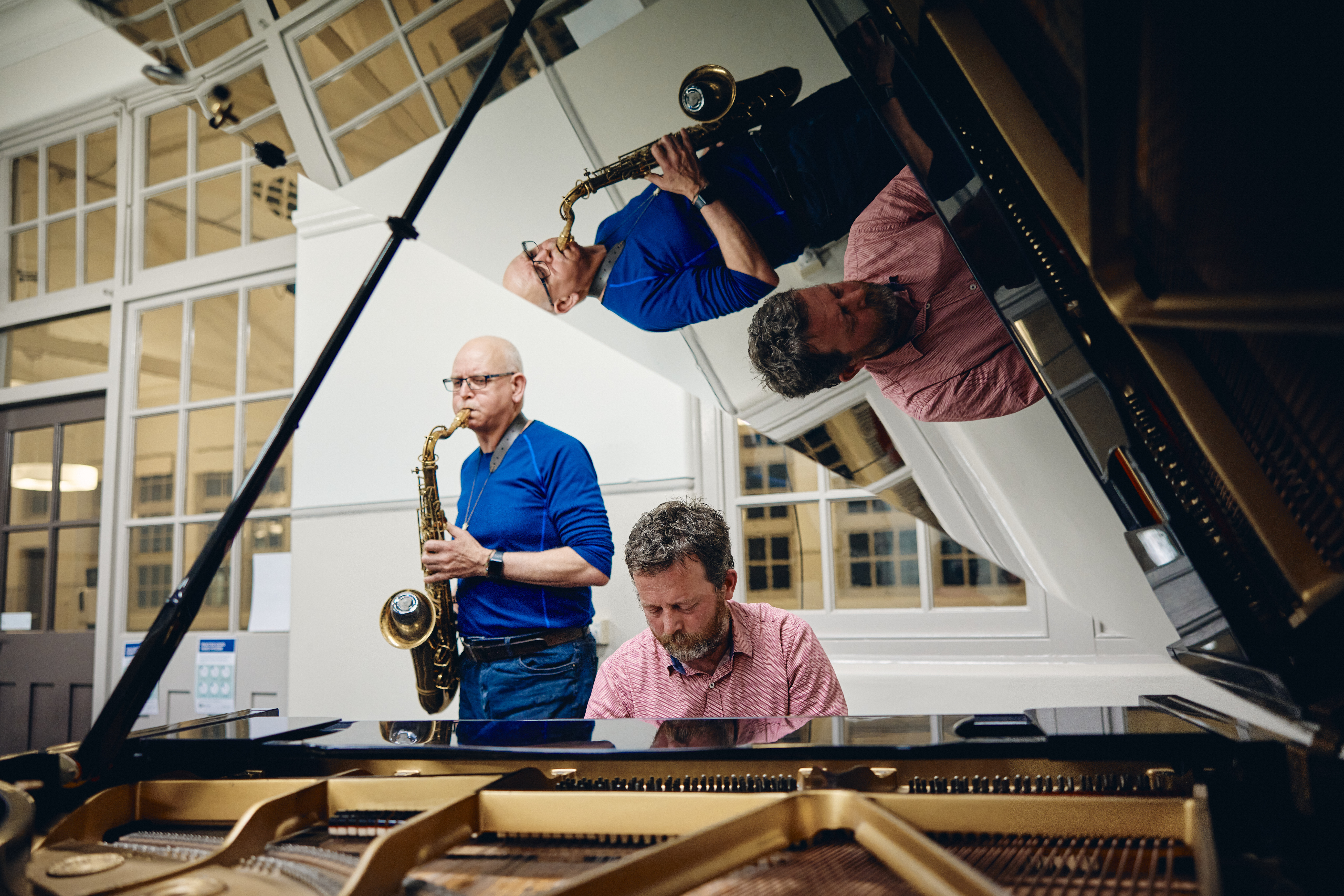 A older man in a red shirt plays a grand piano with a man in a blue shirt playing the saxophone. They are reflected in the open lid of the piano.