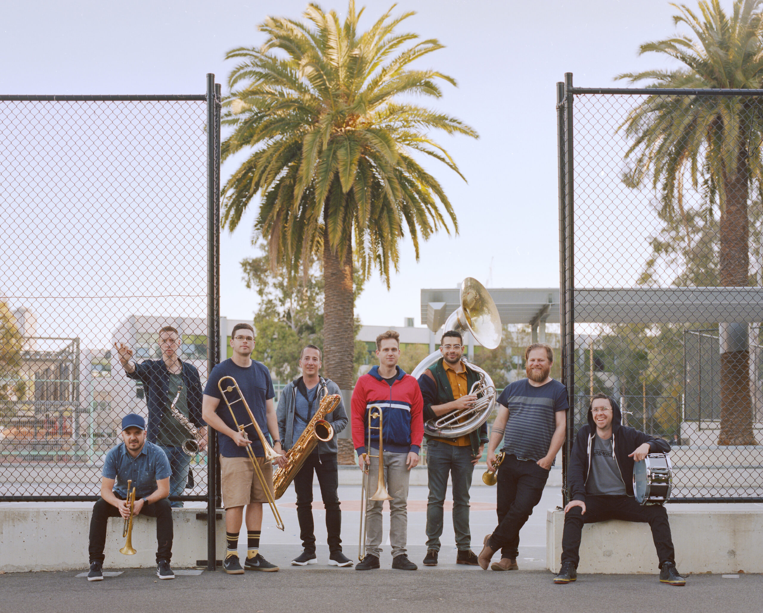 eight musicians holding brass instruments so against a chain linked fence that surrounds a basketball court. Palm trees tower int he background.