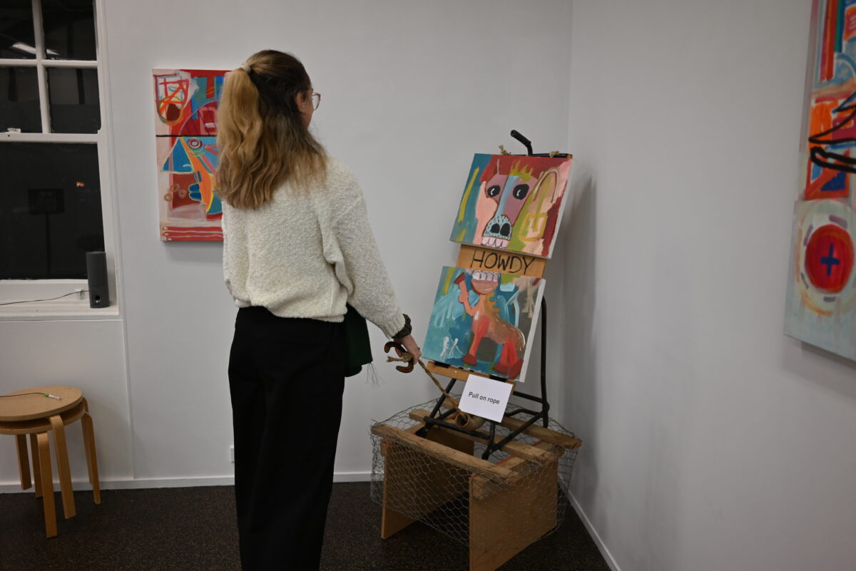A person with long blond hair stands facing towards a colourful series of paintings