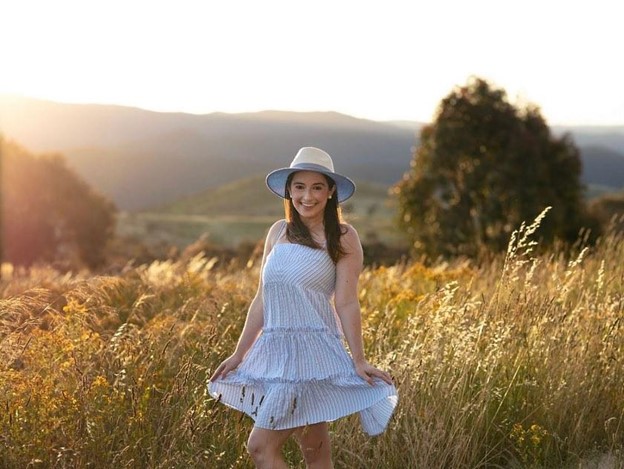 Becky stands in a grassy field with the sun setting behind her. She wears a white dress with a broad-brimmed hat.
