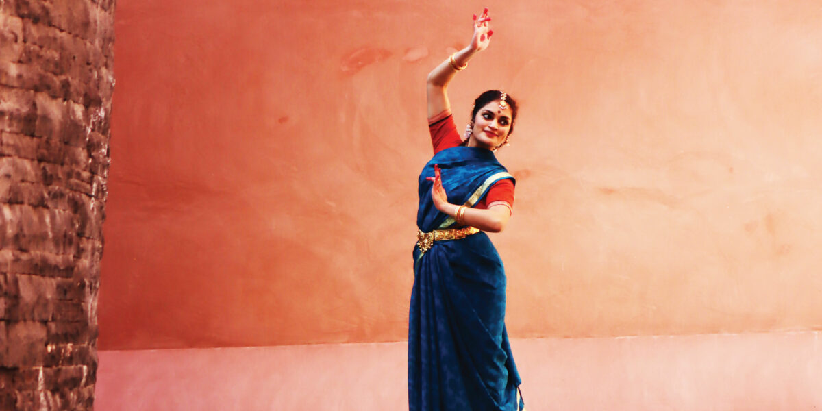 Vaidehi is dressed in a blue and red sari and strikes a pose with one hand above her head with fingers elegantly touching, the other hand is poised in front of her.