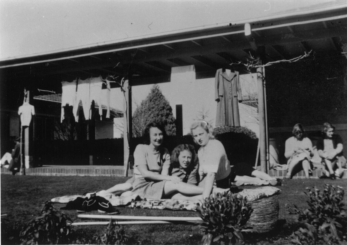 An old black and white photograph. Three women sit on the lawns of Gorman House with their laundry drying in the background.