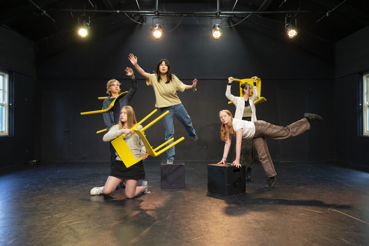 5 young actors pose on a blank stage, as they hold chairs above their heads.