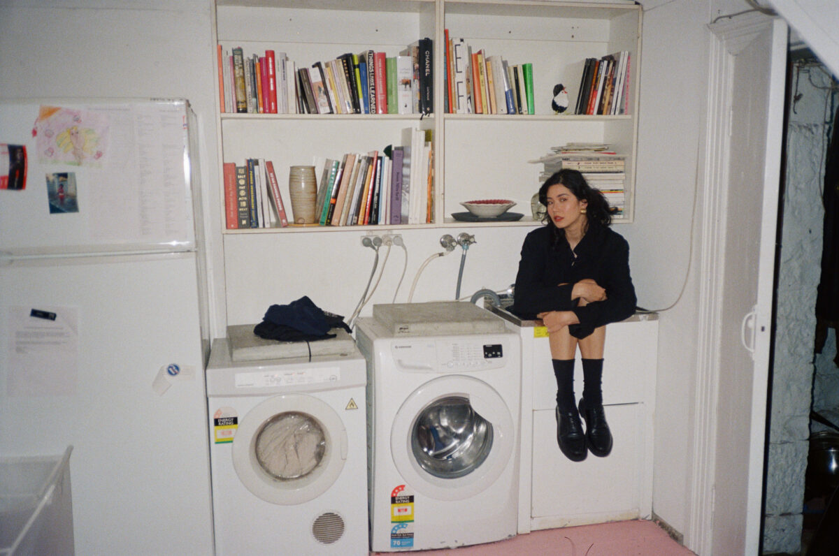 Woman sits in laundry sink with two washer and dryer sitting on the floor. Bookshelves hang on the wall behind her.