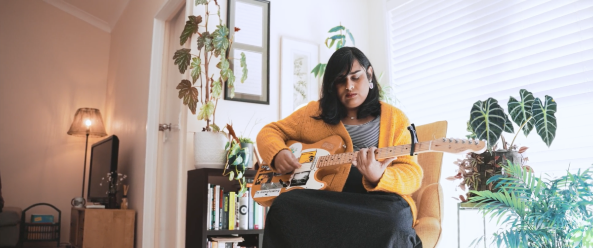 Sia wears a warm yellow cardigan as she strums her electric guitar in her loungeroom. She is sitting down on a chair with a hanging plant and book shelves behind her.