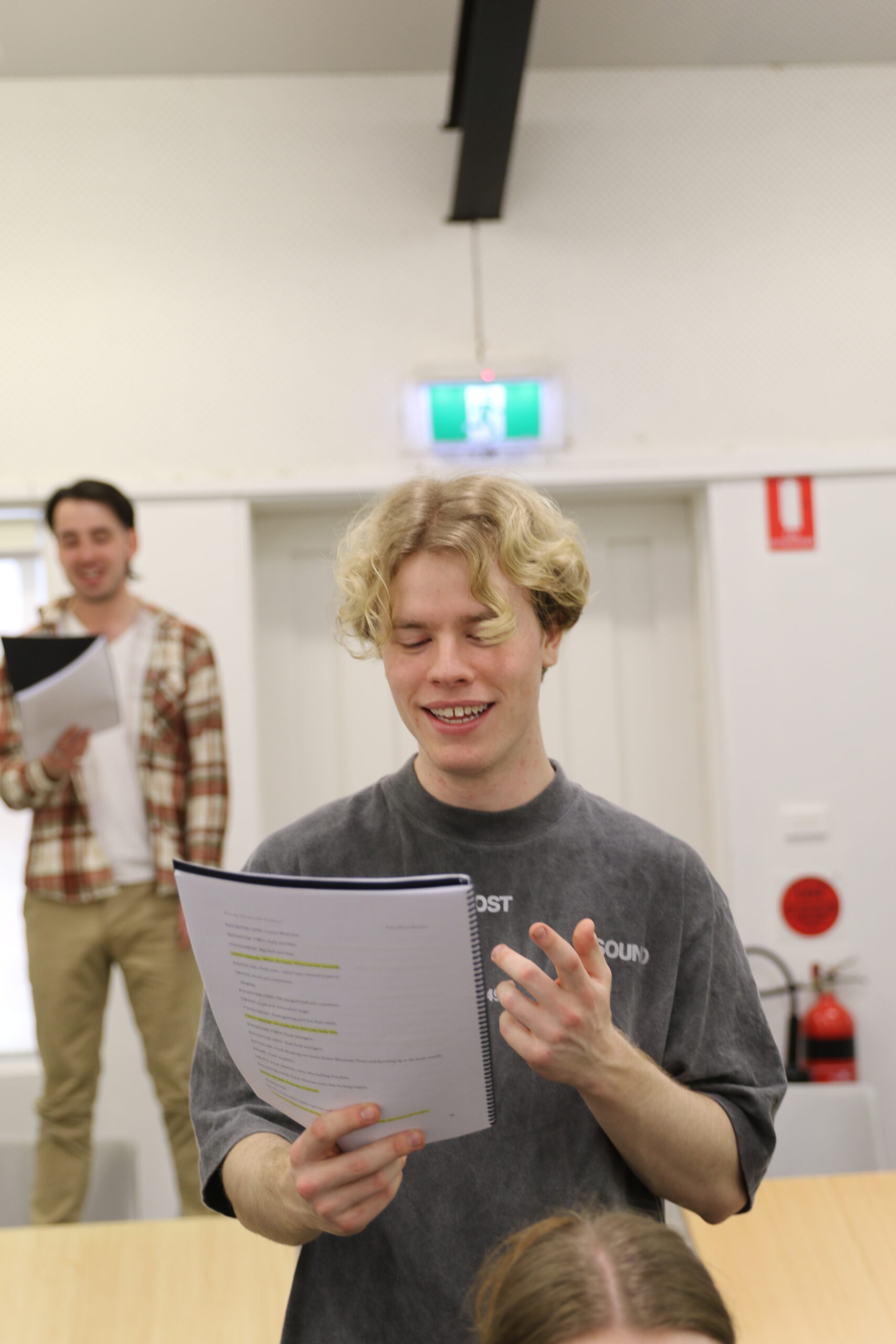 A young actor with cropped blond hair stands reading aloud from a script in from of them.