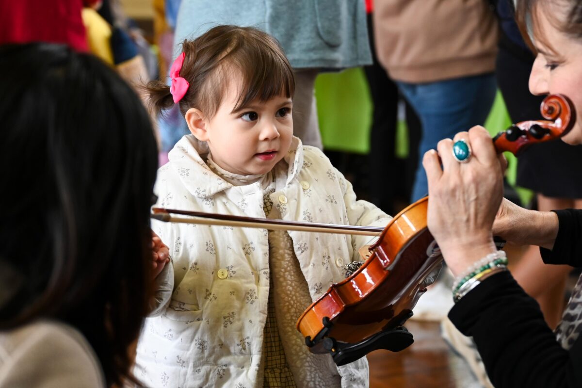 A toddler looks at a violin being held by an adult violinist.