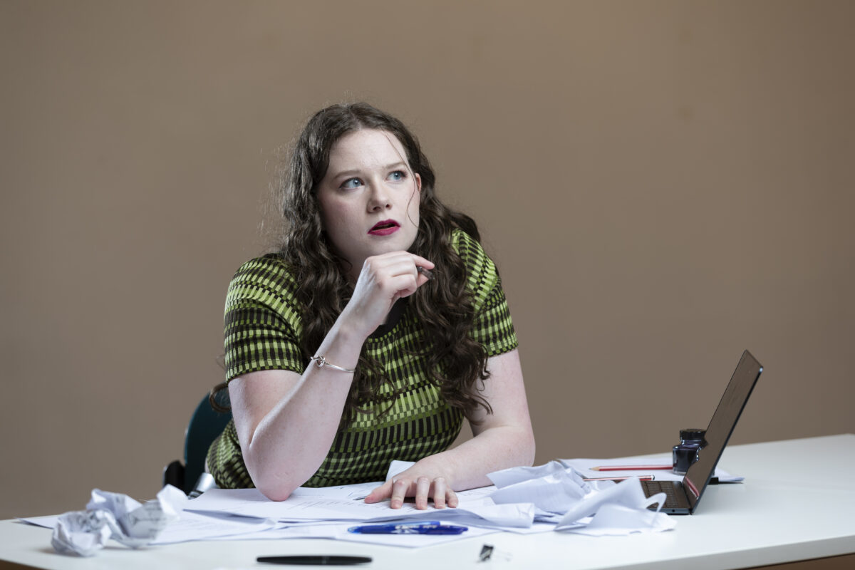 3019_CYT2023_507 – Christopher Carroll A young woman in a green and black shirt sits at a table strewn with papers.