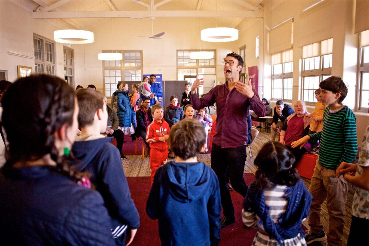The reception hall at Ainslie Arts Centre is filled with children singing and a male conductor standing in front of them, all surrounded by parents and onlookers.