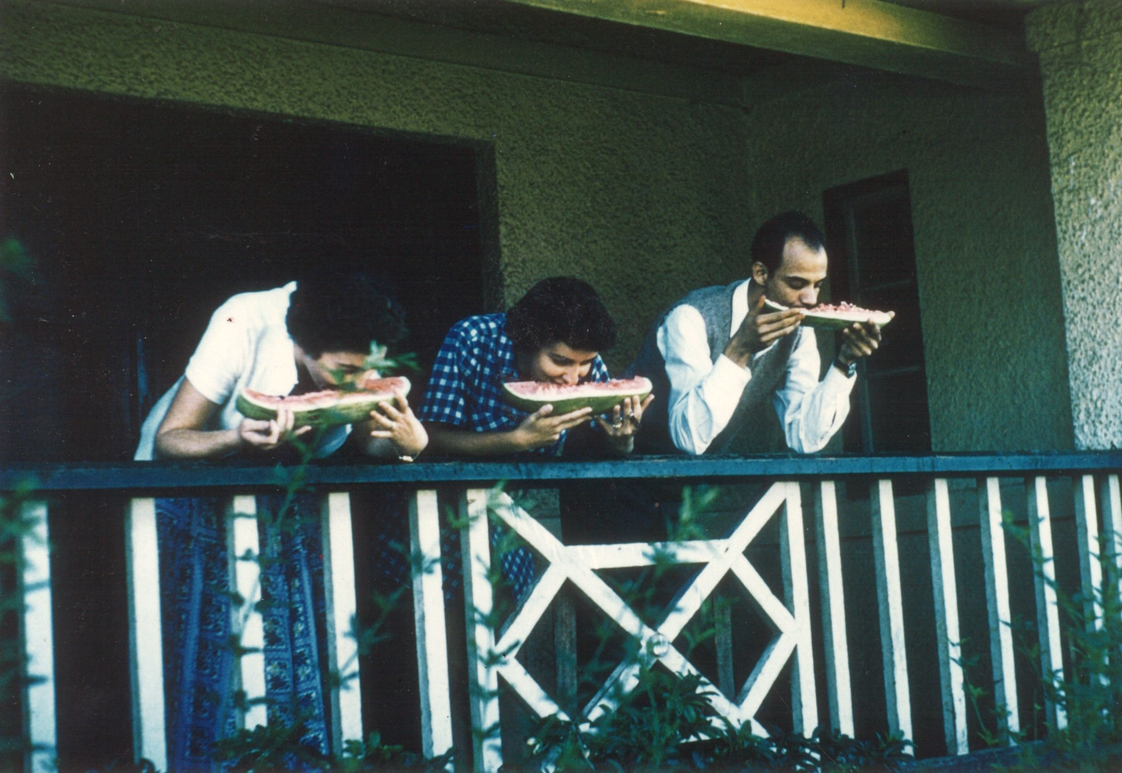 Two women and one man stand on an outdoor veranda eating watermelon.