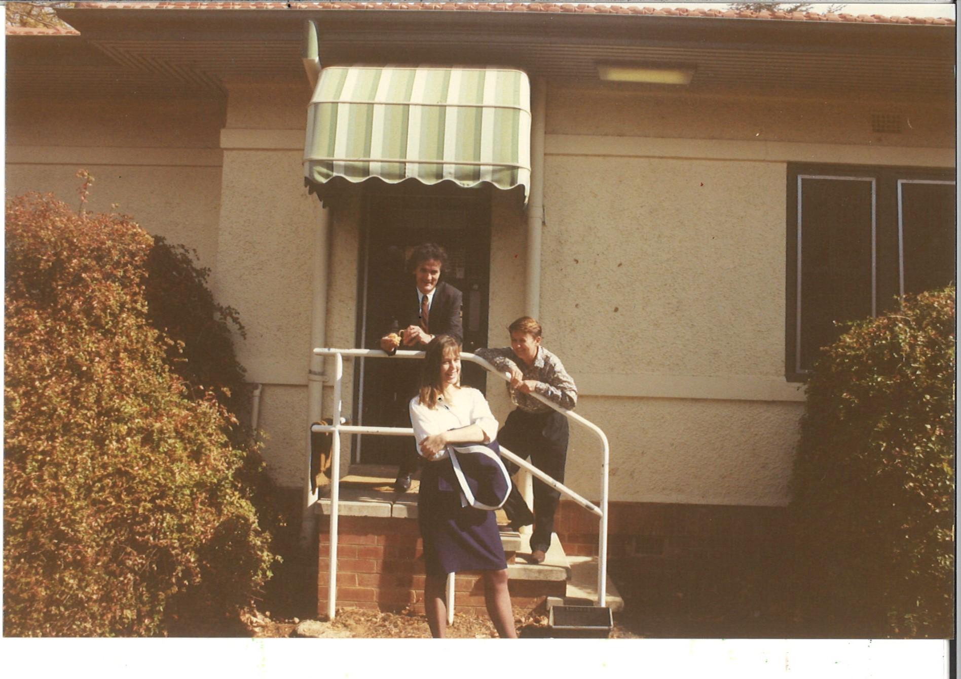 Three people stand on the steps on an old building underneath a striped green and white awning.