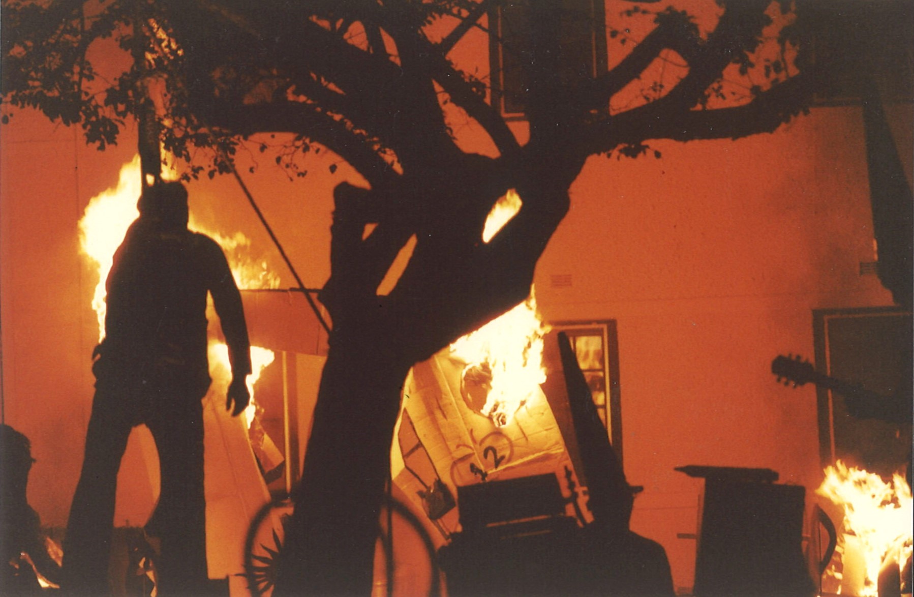 A silhouetted tree lit by firelight with burning stage sets and a figure hanging from a tree.