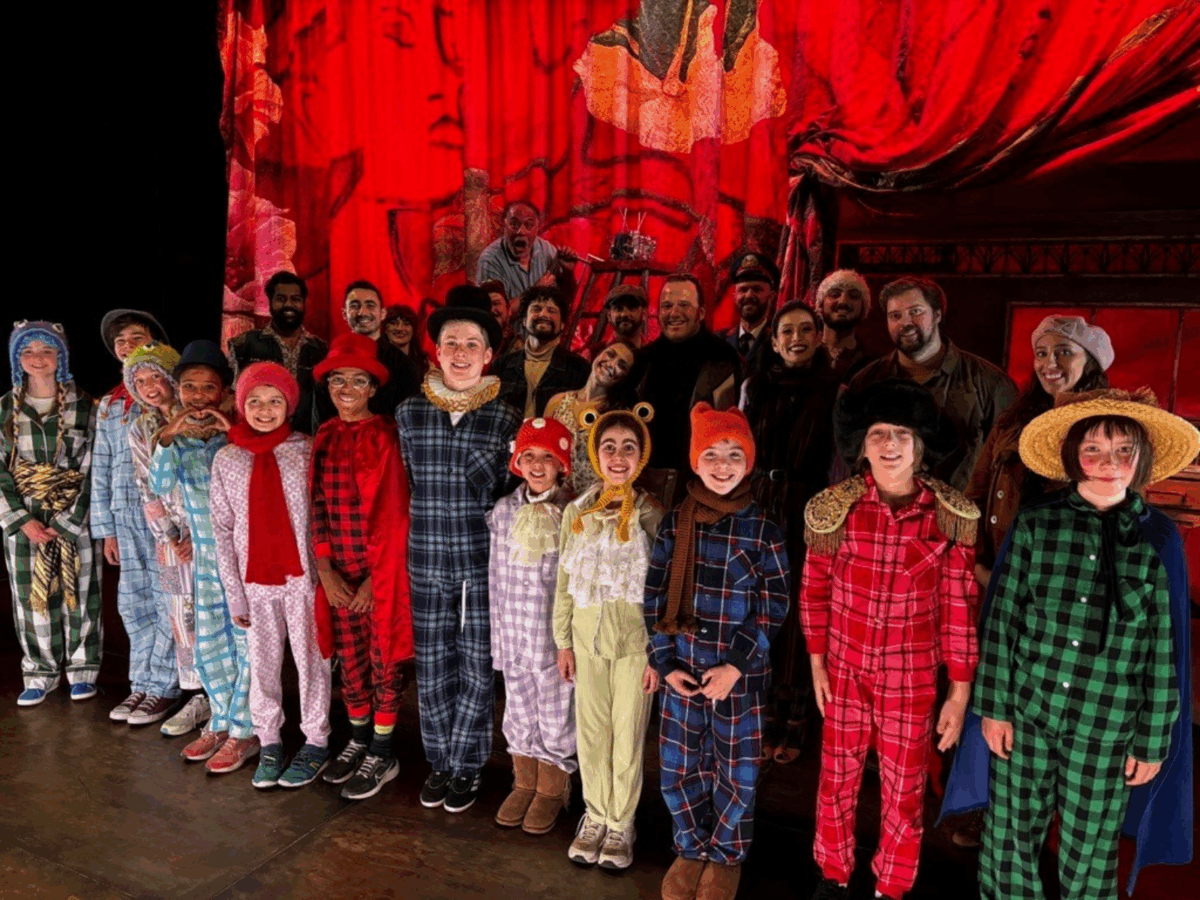Children of all ages stand on a theatre stage in brightly colour costumes smiling at the camera