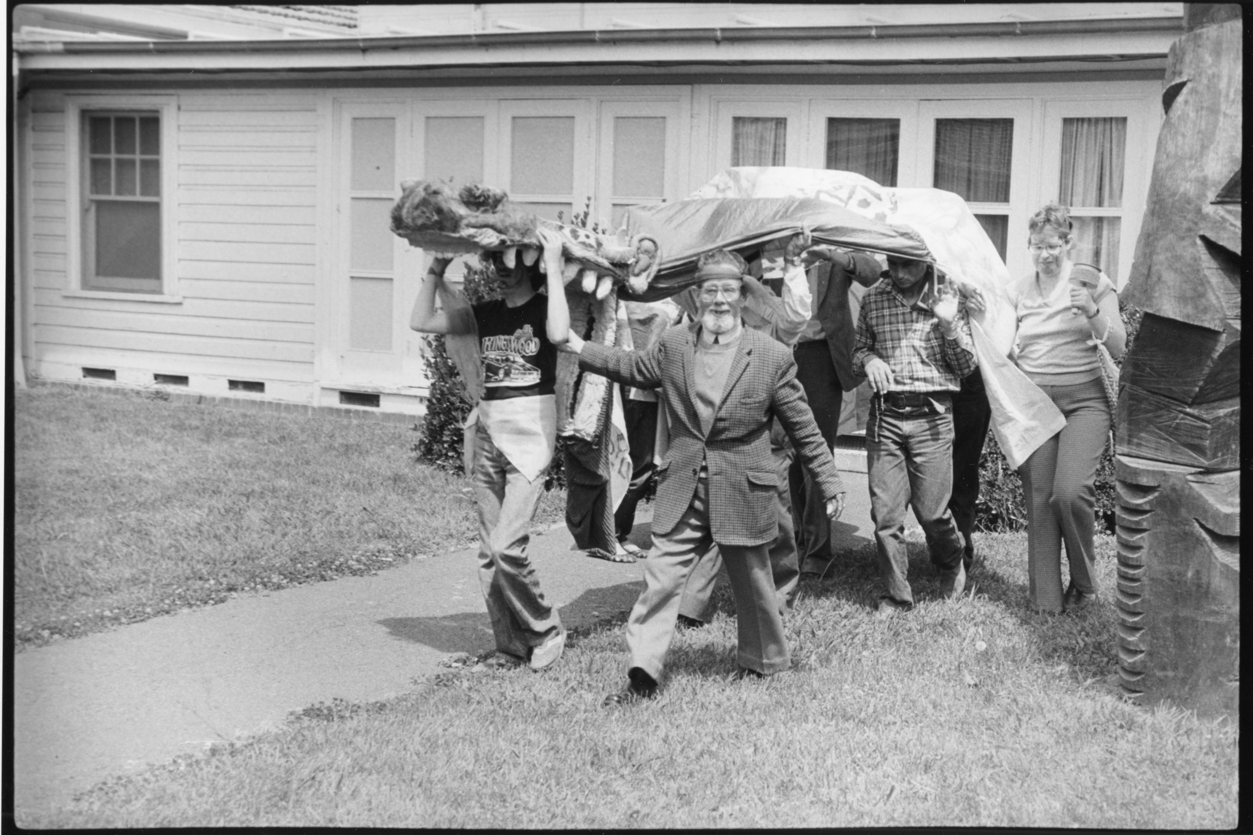A parade of people carrying a sculpture covered in a sheet through the lawns of Gorman House.
