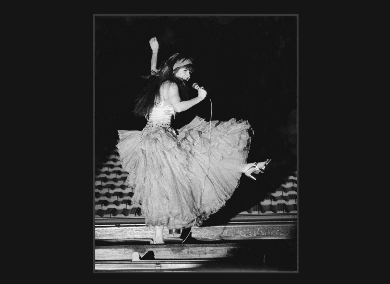 A woman dances on the roof in a long tulle tutu and sings into a microphone.