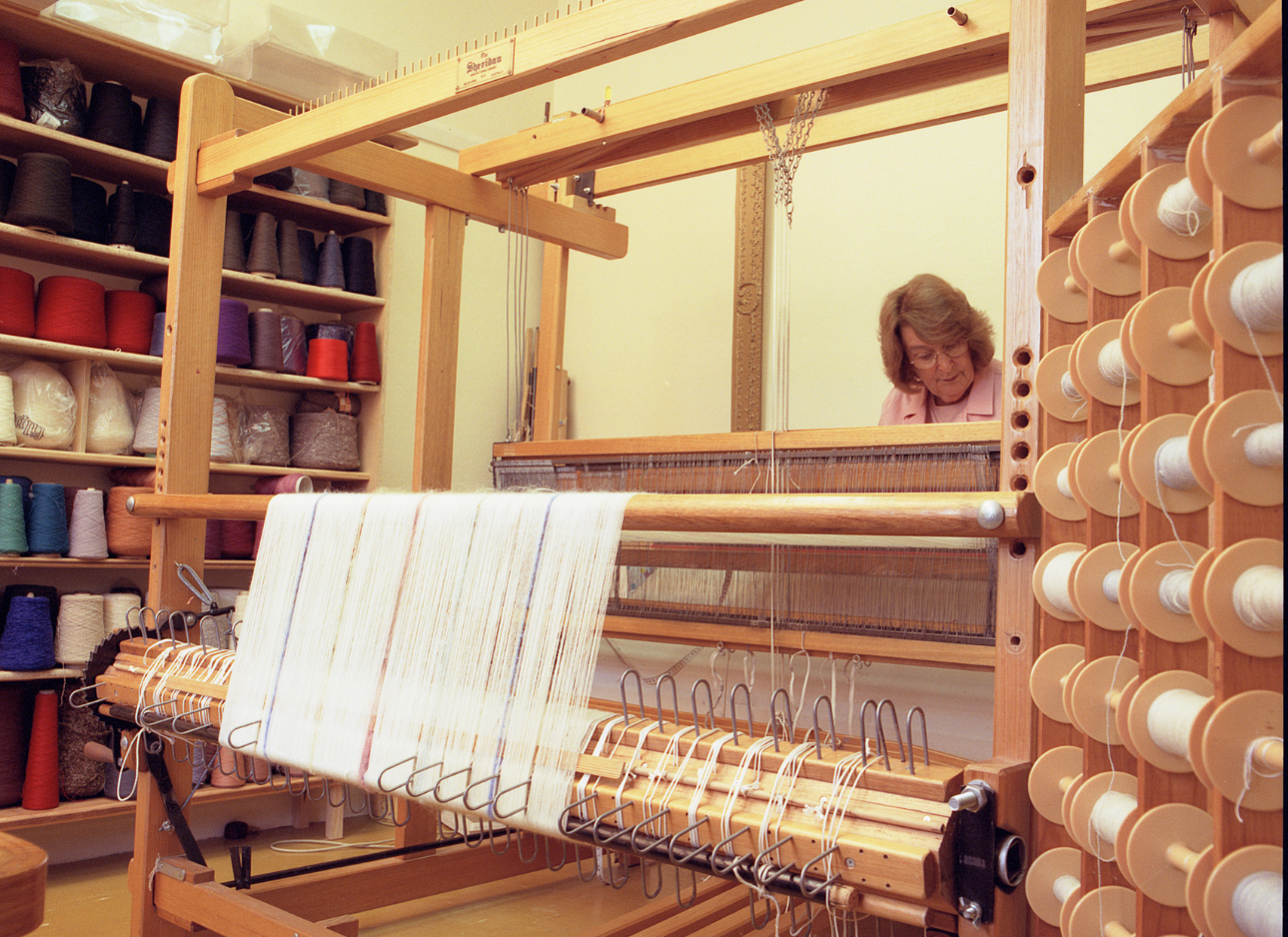 An older woman works at a large weaving loom with white thread spools lining the sides.