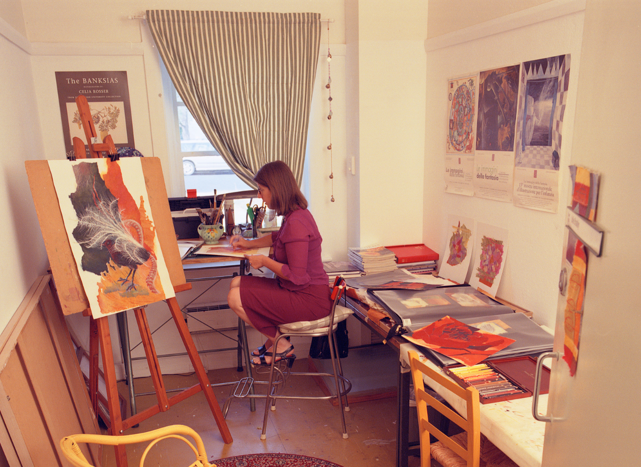 A woman in a red dress, painter Julie Bradley sits at her studio easel next to the window.