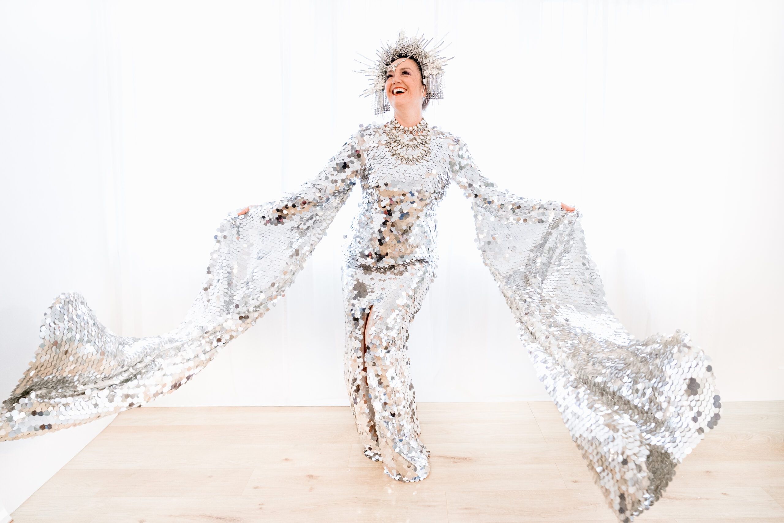 Liz Lea stands triumphantly in a long-sleeved sequinned silver gown with a matching silver crown on a white background and light wooden floor.